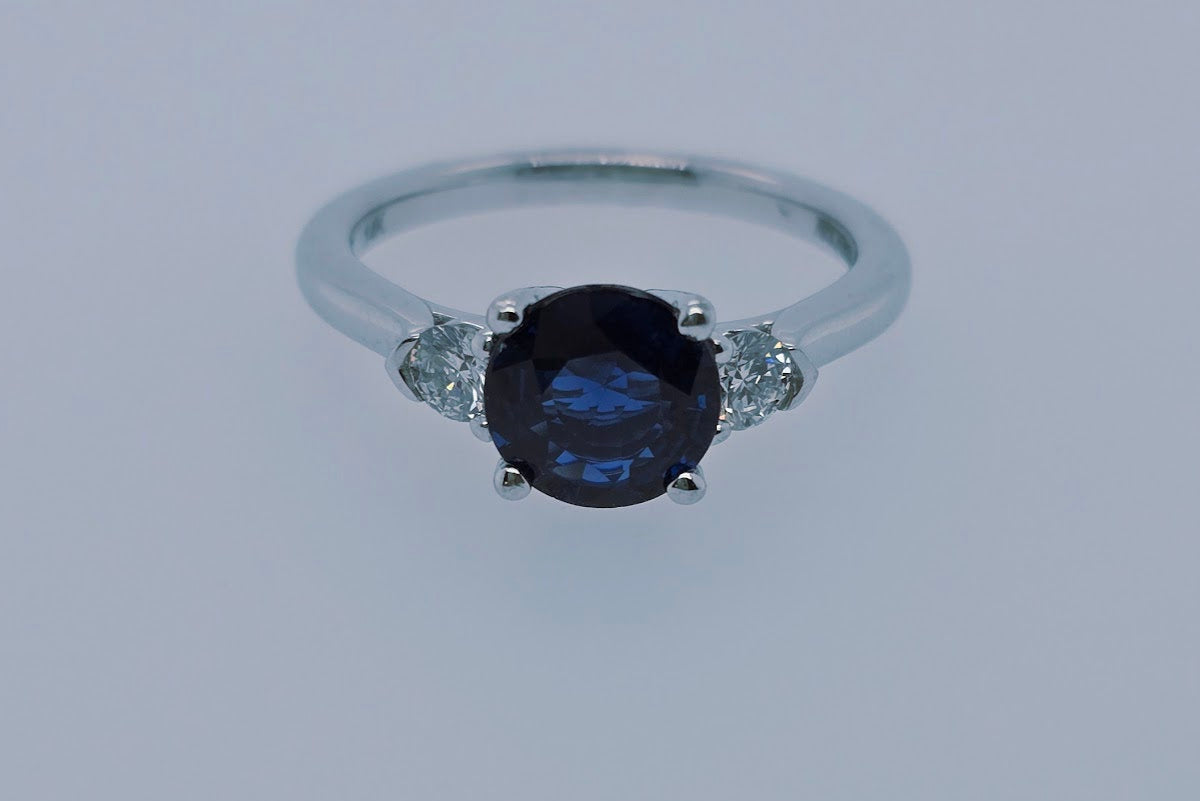 A 14k white gold ring with a deep blue round sapphire center and two sparkling round diamonds, symbolizing past, present, and future.