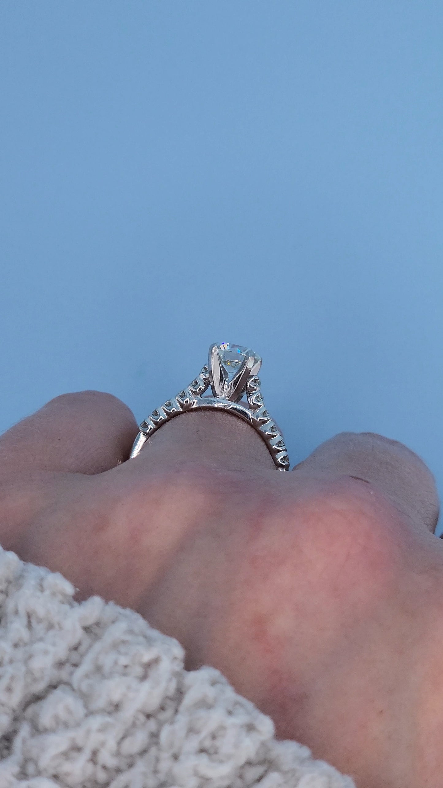 Side view of a white gold engagement ring with a diamond on a finger against a blue background.