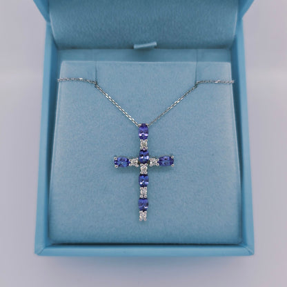 Close-up image of a 14k white gold cross pendant by Effy featuring six vivid oval-cut tanzanite gemstones and six round natural diamonds. The pendant has a hidden bail and hangs from an 18-inch diamond-cut white gold cable chain, displayed on a soft blue background