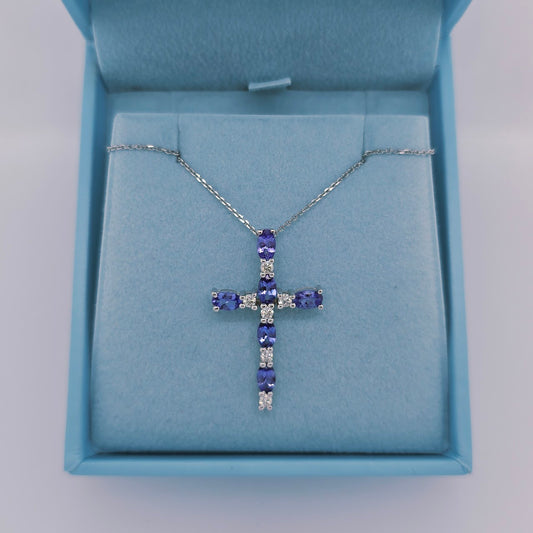 Close-up image of a 14k white gold cross pendant by Effy featuring six vivid oval-cut tanzanite gemstones and six round natural diamonds. The pendant has a hidden bail and hangs from an 18-inch diamond-cut white gold cable chain, displayed on a soft blue background