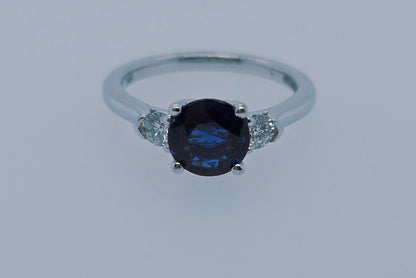A 14k white gold ring with a deep blue round sapphire center and two sparkling round diamonds, symbolizing past, present, and future.